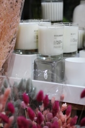 A collection of white candles in glass jars is displayed, with varied labels such as 'Tuileries' and 'Kyoto.' The foreground features a variety of dried, colorful flowers wrapped in clear plastic. The scene conveys a calming yet sophisticated ambiance with its gentle pastel hues and elegant arrangement.