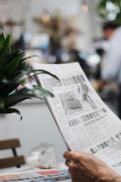 A person is holding a newspaper in a relaxed setting, possibly a cafe or similar environment. The focus is on reading the newspaper, with blurred background elements indicating a busy or casual atmosphere. Plants partially frame the scene, adding a touch of greenery.