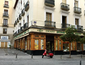 A corner building with a store at street level, featuring large windows and a wooden facade. The words 'Encajes Antonio' are prominently displayed above the shop. Balconies with decorative railings adorn the upper floors, while a red scooter is parked in front. There are some trees lining the street, and a nearby sign advertises the buying and selling of gold.