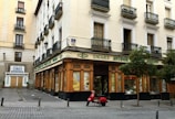 A corner building with a store at street level, featuring large windows and a wooden facade. The words 'Encajes Antonio' are prominently displayed above the shop. Balconies with decorative railings adorn the upper floors, while a red scooter is parked in front. There are some trees lining the street, and a nearby sign advertises the buying and selling of gold.