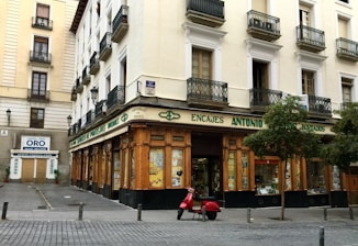 A corner building with a store at street level, featuring large windows and a wooden facade. The words 'Encajes Antonio' are prominently displayed above the shop. Balconies with decorative railings adorn the upper floors, while a red scooter is parked in front. There are some trees lining the street, and a nearby sign advertises the buying and selling of gold.