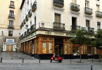 A corner building with a store at street level, featuring large windows and a wooden facade. The words 'Encajes Antonio' are prominently displayed above the shop. Balconies with decorative railings adorn the upper floors, while a red scooter is parked in front. There are some trees lining the street, and a nearby sign advertises the buying and selling of gold.