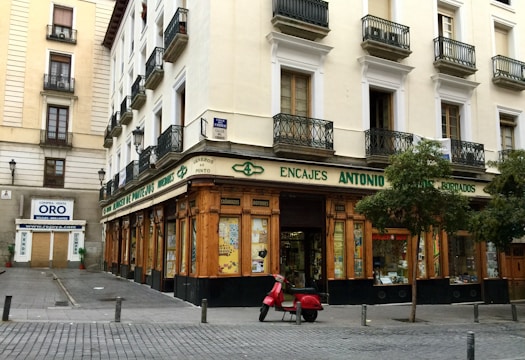 A corner building with a store at street level, featuring large windows and a wooden facade. The words 'Encajes Antonio' are prominently displayed above the shop. Balconies with decorative railings adorn the upper floors, while a red scooter is parked in front. There are some trees lining the street, and a nearby sign advertises the buying and selling of gold.