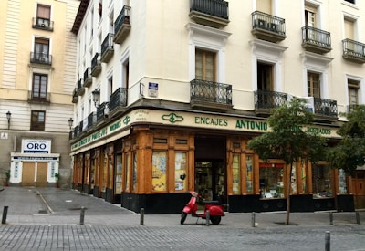 A corner building with a store at street level, featuring large windows and a wooden facade. The words 'Encajes Antonio' are prominently displayed above the shop. Balconies with decorative railings adorn the upper floors, while a red scooter is parked in front. There are some trees lining the street, and a nearby sign advertises the buying and selling of gold.