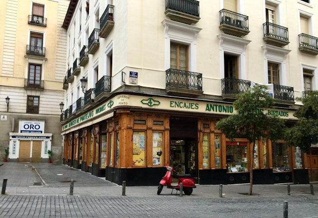 A corner building with a store at street level, featuring large windows and a wooden facade. The words 'Encajes Antonio' are prominently displayed above the shop. Balconies with decorative railings adorn the upper floors, while a red scooter is parked in front. There are some trees lining the street, and a nearby sign advertises the buying and selling of gold.