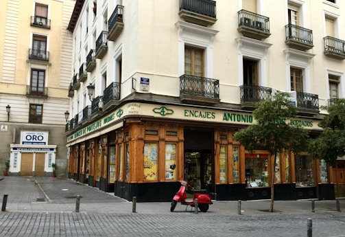 A corner building with a store at street level, featuring large windows and a wooden facade. The words 'Encajes Antonio' are prominently displayed above the shop. Balconies with decorative railings adorn the upper floors, while a red scooter is parked in front. There are some trees lining the street, and a nearby sign advertises the buying and selling of gold.