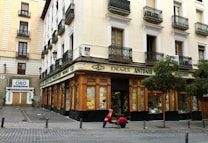A corner building with a store at street level, featuring large windows and a wooden facade. The words 'Encajes Antonio' are prominently displayed above the shop. Balconies with decorative railings adorn the upper floors, while a red scooter is parked in front. There are some trees lining the street, and a nearby sign advertises the buying and selling of gold.