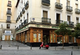 A corner building with a store at street level, featuring large windows and a wooden facade. The words 'Encajes Antonio' are prominently displayed above the shop. Balconies with decorative railings adorn the upper floors, while a red scooter is parked in front. There are some trees lining the street, and a nearby sign advertises the buying and selling of gold.