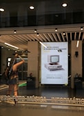 A storefront with a modern design features large windows and a bright interior. A life-sized statue of a warrior is positioned near the entrance, holding weapons and wearing armor. Inside, a poster displays an old-fashioned computer, suggesting updates or technological advancements. The text on the poster is in Spanish, advertising electronic products. Overhead lights illuminate the scene, and reflections are visible on the glass.