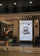 A storefront with a modern design features large windows and a bright interior. A life-sized statue of a warrior is positioned near the entrance, holding weapons and wearing armor. Inside, a poster displays an old-fashioned computer, suggesting updates or technological advancements. The text on the poster is in Spanish, advertising electronic products. Overhead lights illuminate the scene, and reflections are visible on the glass.