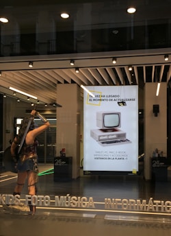 A storefront with a modern design features large windows and a bright interior. A life-sized statue of a warrior is positioned near the entrance, holding weapons and wearing armor. Inside, a poster displays an old-fashioned computer, suggesting updates or technological advancements. The text on the poster is in Spanish, advertising electronic products. Overhead lights illuminate the scene, and reflections are visible on the glass.