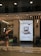 A storefront with a modern design features large windows and a bright interior. A life-sized statue of a warrior is positioned near the entrance, holding weapons and wearing armor. Inside, a poster displays an old-fashioned computer, suggesting updates or technological advancements. The text on the poster is in Spanish, advertising electronic products. Overhead lights illuminate the scene, and reflections are visible on the glass.