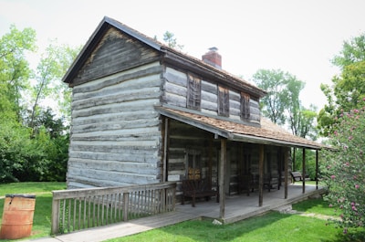 A rustic log cabin with a weathered wood exterior, featuring a front porch with wooden chairs. The cabin's roof has a slight pitch, with a small chimney protruding from the center. The surrounding area is lush with green grass, trees, and shrubs, providing a serene and natural setting.
