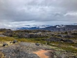A rugged, rocky landscape with patches of green and yellow moss, stretching towards distant mountains shrouded in mist. The sky is overcast with heavy grey clouds, contributing to a moody and atmospheric scene.