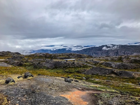 A rugged, rocky landscape with patches of green and yellow moss, stretching towards distant mountains shrouded in mist. The sky is overcast with heavy grey clouds, contributing to a moody and atmospheric scene.