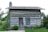 A rustic log cabin with horizontal wooden beams and a stone chimney stands amidst a lush garden. The cabin features a wooden door and a closed wooden shutter. Surrounding the cabin are green shrubs and flowers, providing a natural setting.