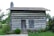 A rustic log cabin with horizontal wooden beams and a stone chimney stands amidst a lush garden. The cabin features a wooden door and a closed wooden shutter. Surrounding the cabin are green shrubs and flowers, providing a natural setting.