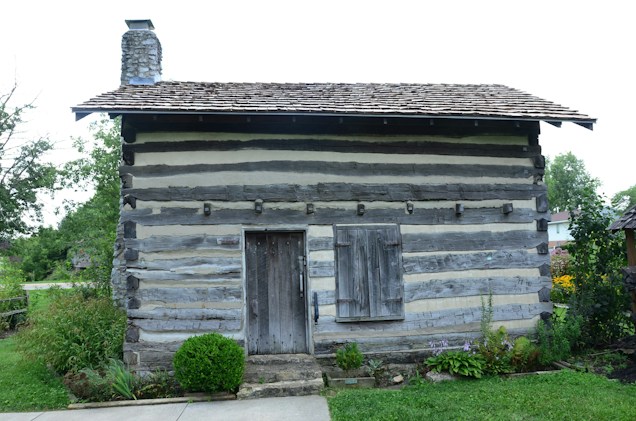 A rustic log cabin with horizontal wooden beams and a stone chimney stands amidst a lush garden. The cabin features a wooden door and a closed wooden shutter. Surrounding the cabin are green shrubs and flowers, providing a natural setting.