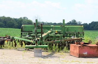 An old, green agricultural machine sits in a rural field, surrounded by lush greenery and trees in the distance. The machine appears to be weathered and unused, positioned on a small patch of concrete. A rust-colored box lies nearby, adding contrast to the scene.