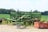 An old, green agricultural machine sits in a rural field, surrounded by lush greenery and trees in the distance. The machine appears to be weathered and unused, positioned on a small patch of concrete. A rust-colored box lies nearby, adding contrast to the scene.
