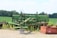 An old, green agricultural machine sits in a rural field, surrounded by lush greenery and trees in the distance. The machine appears to be weathered and unused, positioned on a small patch of concrete. A rust-colored box lies nearby, adding contrast to the scene.