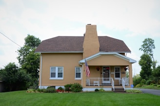 A small, well-maintained house with a brown roof, yellow exterior, and white trim is shown. The house has a front porch with white railings, potted plants, a few outdoor chairs, and an American flag hanging. The front yard is lush with green grass, and there are various bushes and plants along the base of the house. Tall trees and some shrubs can be seen in the background along with a clear blue sky.