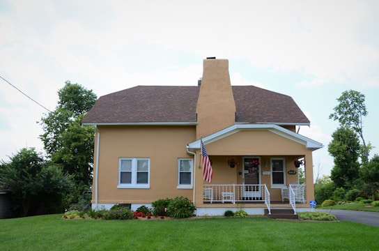 A small, well-maintained house with a brown roof, yellow exterior, and white trim is shown. The house has a front porch with white railings, potted plants, a few outdoor chairs, and an American flag hanging. The front yard is lush with green grass, and there are various bushes and plants along the base of the house. Tall trees and some shrubs can be seen in the background along with a clear blue sky.
