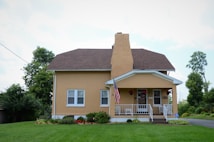 A small, well-maintained house with a brown roof, yellow exterior, and white trim is shown. The house has a front porch with white railings, potted plants, a few outdoor chairs, and an American flag hanging. The front yard is lush with green grass, and there are various bushes and plants along the base of the house. Tall trees and some shrubs can be seen in the background along with a clear blue sky.