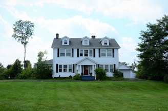 A large, two-story colonial-style house with white siding and black shutters, surrounded by lush greenery. The front of the house features a small porch with red steps and several windows. The lawn in front is well-manicured, and there are tall trees and bushes around the perimeter, set against a partly cloudy sky.