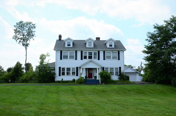 A charming two-story colonial home with a bright red door and manicured front lawn under a clear blue sky.