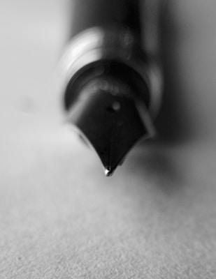 A black and white close-up of a fountain pen nib, with a soft focus that emphasizes the sharp tip and intricate design of the nib. The pen rests on a textured surface, creating a minimalistic and elegant aesthetic.