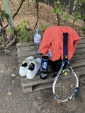 A tennis racket lies on a wooden bench alongside a pair of white tennis shoes with green accents. A blue canister, possibly for tennis balls, is next to an orange shirt draped over a black bag. A large bottle of water sits nearby, with a wire fence and greenery in the background.