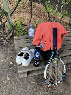 A tennis racket lies on a wooden bench alongside a pair of white tennis shoes with green accents. A blue canister, possibly for tennis balls, is next to an orange shirt draped over a black bag. A large bottle of water sits nearby, with a wire fence and greenery in the background.