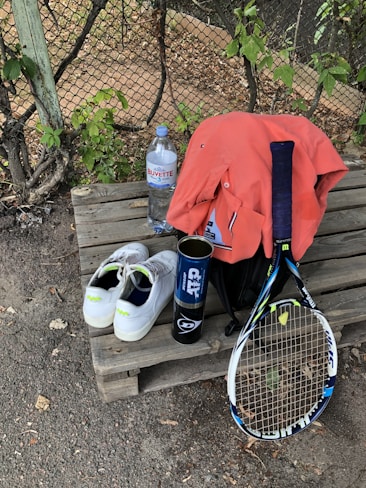 A tennis racket lies on a wooden bench alongside a pair of white tennis shoes with green accents. A blue canister, possibly for tennis balls, is next to an orange shirt draped over a black bag. A large bottle of water sits nearby, with a wire fence and greenery in the background.