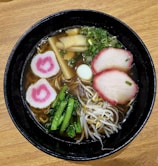 A bowl of ramen features a rich broth with noodles, topped with slices of pink-edged meat, spiral-shaped fish cakes with a pink swirl, bean sprouts, green onions, bamboo shoots, and leafy greens. The setting has a wooden table surface.