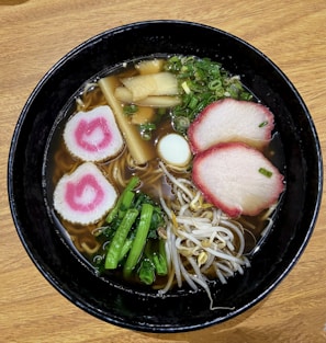A bowl of ramen features a rich broth with noodles, topped with slices of pink-edged meat, spiral-shaped fish cakes with a pink swirl, bean sprouts, green onions, bamboo shoots, and leafy greens. The setting has a wooden table surface.