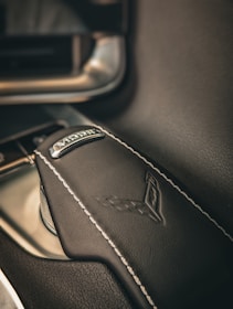 A close-up shot of a car's gear lever, wrapped in black leather with white stitching. The lever features a metallic emblem and a stitched logo of a sports or luxury car brand.