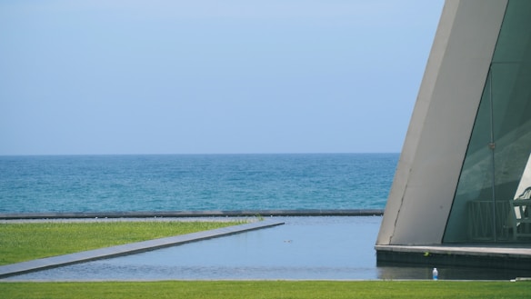 A tranquil seascape with a clear blue sky and calm ocean, featuring a minimalist modern structure with large glass windows and a sharp triangular form. The foreground includes a well-maintained grass lawn and a reflective water feature, enhancing the serene atmosphere.