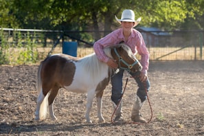 A person wearing a cowboy hat and striped shirt is standing outside on a dirt ground, holding a small pony by the bridle. The pony has a brown and white coat and is adorned with a turquoise halter. The background features a fence, trees, and a blurred structure in the distance, creating a rural setting.