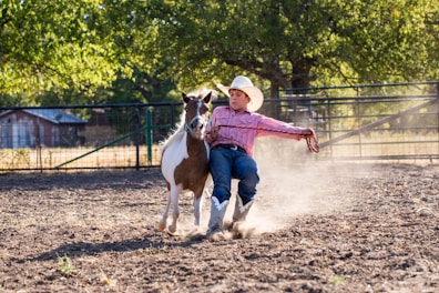 In a dusty outdoor setting, a young person wearing a cowboy hat, a striped shirt, blue jeans, and boots is seen leading a small horse by the reins. The person is leaning back, creating a sense of motion and action.