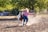 In a dusty outdoor setting, a young person wearing a cowboy hat, a striped shirt, blue jeans, and boots is seen leading a small horse by the reins. The person is leaning back, creating a sense of motion and action.
