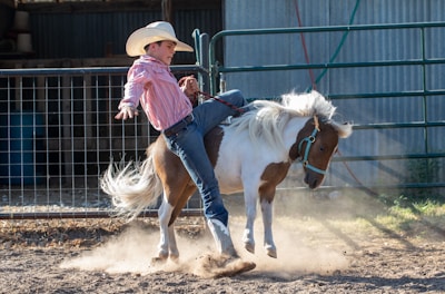 A young person wearing a cowboy hat and a striped shirt rides a small bucking horse in a dusty corral. The scene captures movement and a playful yet challenging moment in an outdoor ranch setting.