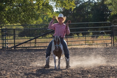 A person wearing a cowboy hat and a striped pink shirt is riding a horse in a dusty outdoor enclosure. The horse is brown and white, and the scene is surrounded by metal fencing and greenery in the background. The rider is holding a rope in one hand.