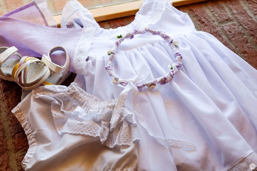 A neatly arranged set of light purple and white clothing and accessories is displayed, including a white dress with lace details, a pair of matching sandals, a headband with fabric flowers, and a pair of bloomers. The ensemble is laid out on a wooden surface with a warm background.