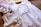A neatly arranged set of light purple and white clothing and accessories is displayed, including a white dress with lace details, a pair of matching sandals, a headband with fabric flowers, and a pair of bloomers. The ensemble is laid out on a wooden surface with a warm background.