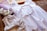 A neatly arranged set of light purple and white clothing and accessories is displayed, including a white dress with lace details, a pair of matching sandals, a headband with fabric flowers, and a pair of bloomers. The ensemble is laid out on a wooden surface with a warm background.