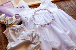 A neatly arranged set of light purple and white clothing and accessories is displayed, including a white dress with lace details, a pair of matching sandals, a headband with fabric flowers, and a pair of bloomers. The ensemble is laid out on a wooden surface with a warm background.