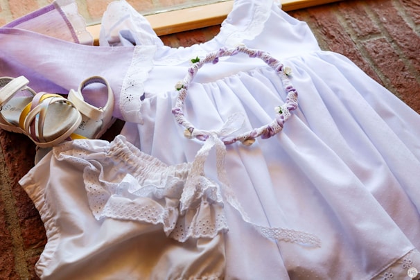 A neatly arranged set of light purple and white clothing and accessories is displayed, including a white dress with lace details, a pair of matching sandals, a headband with fabric flowers, and a pair of bloomers. The ensemble is laid out on a wooden surface with a warm background.