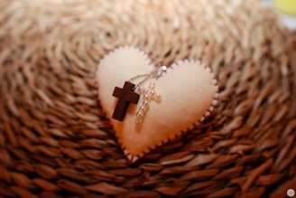 A soft heart-shaped object made of fabric placed on a textured background. On top of the heart lies a wooden cross and a string of pearls forming another cross. The item rests on a woven, circular mat, creating a warm and cozy atmosphere.