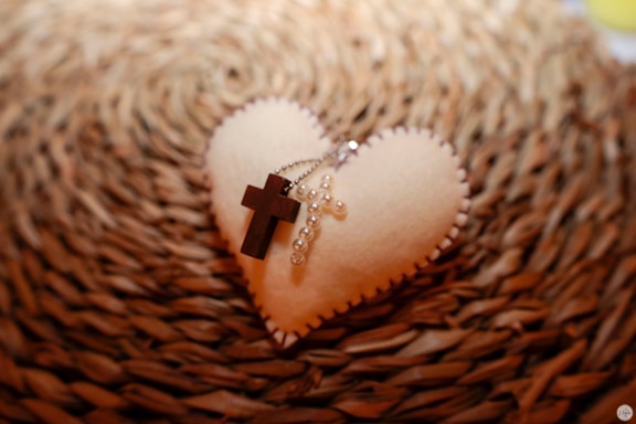 A soft heart-shaped object made of fabric placed on a textured background. On top of the heart lies a wooden cross and a string of pearls forming another cross. The item rests on a woven, circular mat, creating a warm and cozy atmosphere.